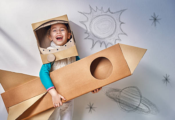 A child holds a cardboard box rocket ship while wearing a cardboard box helmet. A child holds a cardboard box rocket ship while wearing a cardboard box helmet.