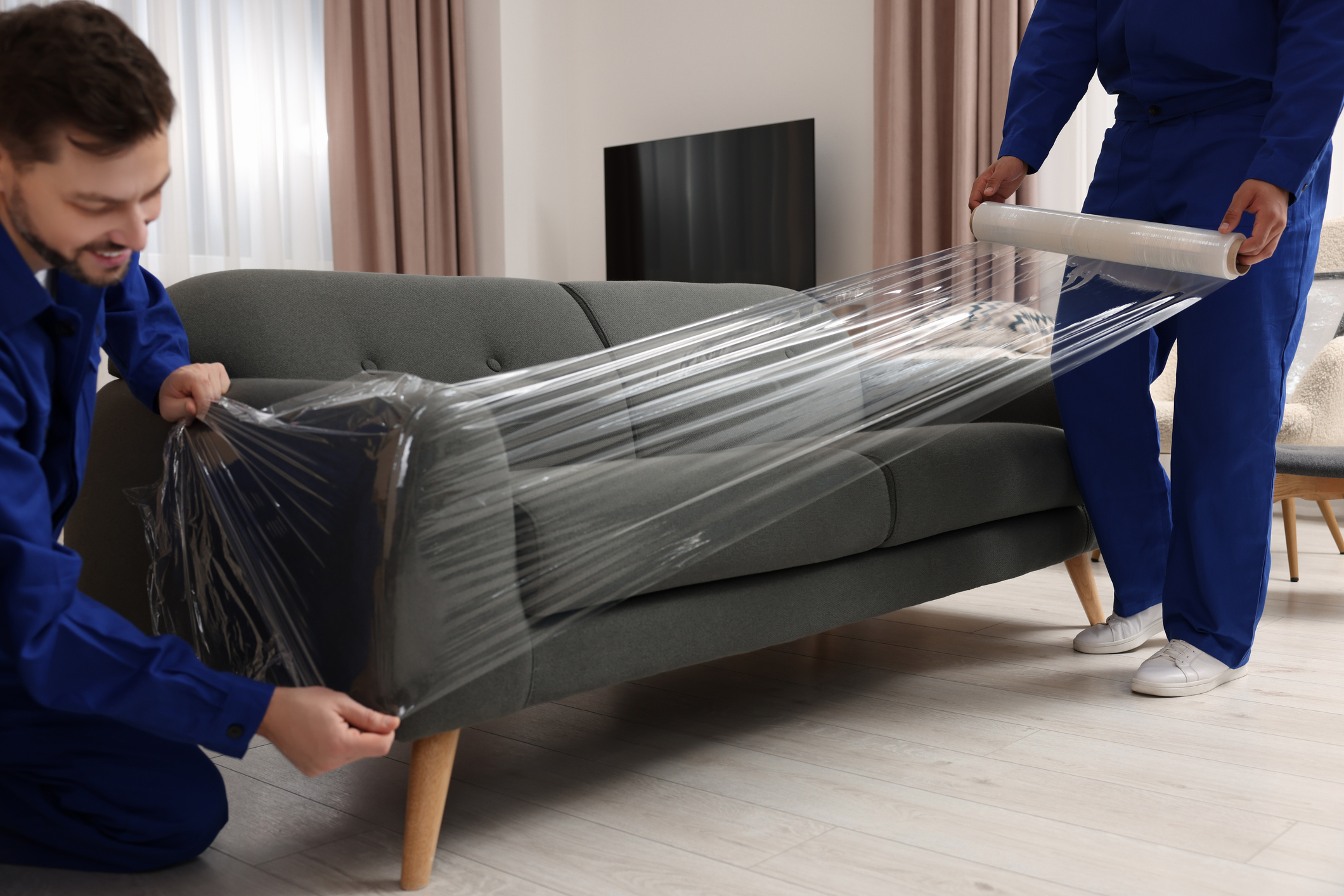 Two movers wrapping a couch in plastic wrap to keep the cushion on. 