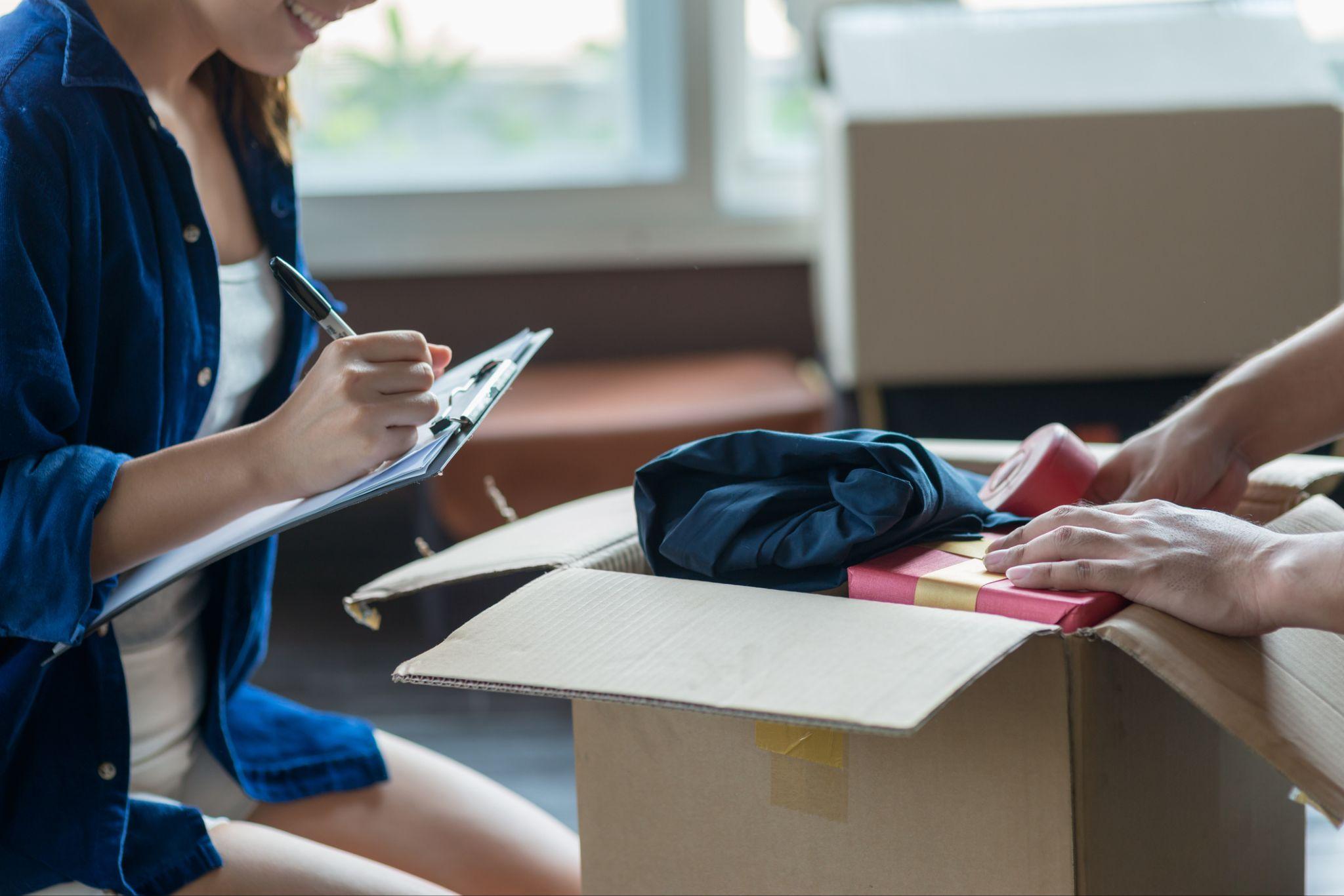 A woman marks off a checklist as someone else packs a moving box next to her.