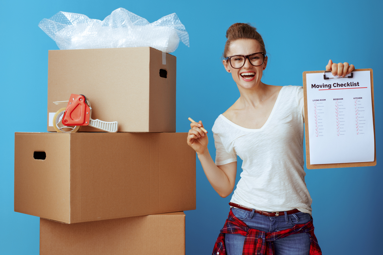 A woman with a checklist stands nest to a tower of boxes and moving supplies.