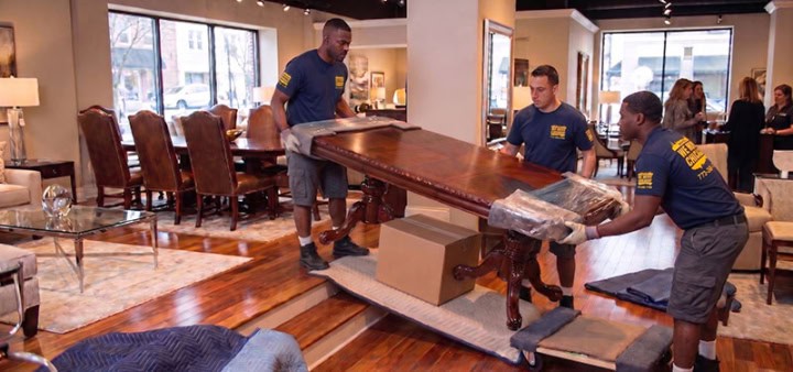 Aaron Bros Moving System crew carrying a wrapped dining table through a furniture showroom